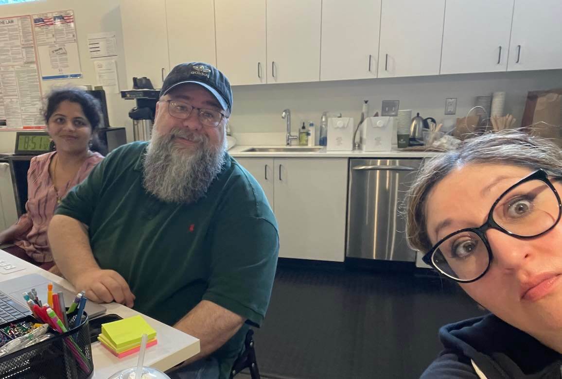 Three people pose together in an office, either making faces or smiling at the camera as one of them takes a selfie